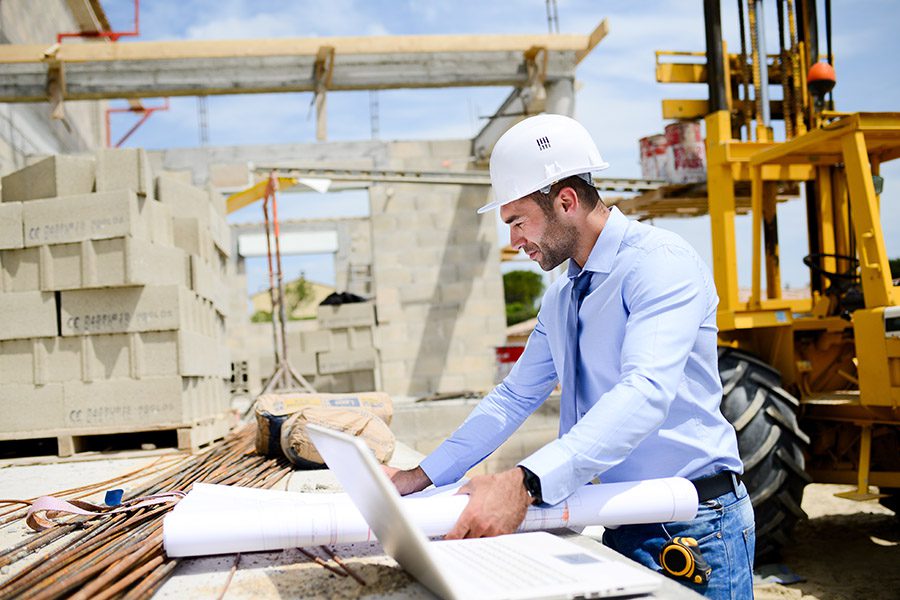 Specialized Business Insurance - An Engineer is Reviewing Project Plans at a Construction Site With Bricks and a Construction Vehicle Behind Him During a Sunny Day